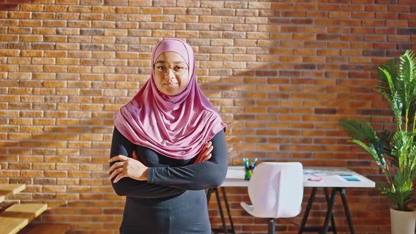 Muslim Woman in a Hijab and Glasses with Folded Hands Stands Near Her Desk in the Office or at Home