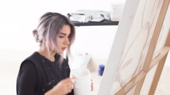 Cheerful Latina Girl Holding Gesso White Paint And A Palette Knife Working On A Canvas. - Medium Sho