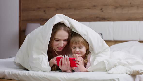 Mother Lying on Bed Under Duvet Blanket with Daughter Kid Girl Holding Mobile Phone Watching Cartoon