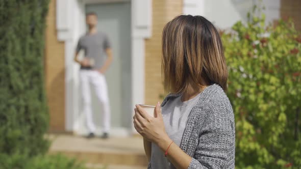 Cute Caucasian Woman Standing with the Cup of Tea in the Foreground, Her Husband Coming From Behind