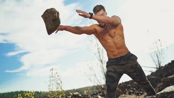 Angry sportsman with muscular body throws stones in nature. Shirtless bodybuilder exercises with hea