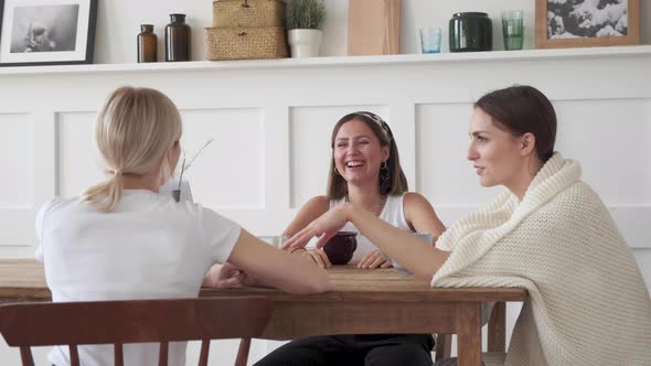 Three girlfriends are smiling sitting at home at the table and talking about a new project