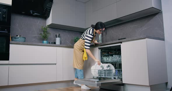 Brunette Housewife in Apron Standing in front of Dishwasher and Putting Wash Dishes