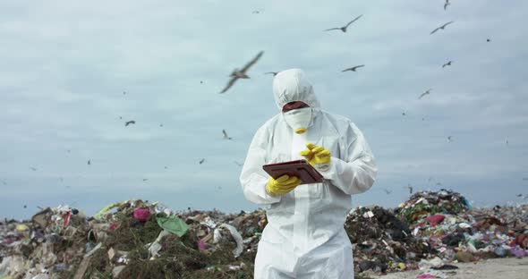 Virologist in Protective Costume Noting in Tablet During Researching a Landfill