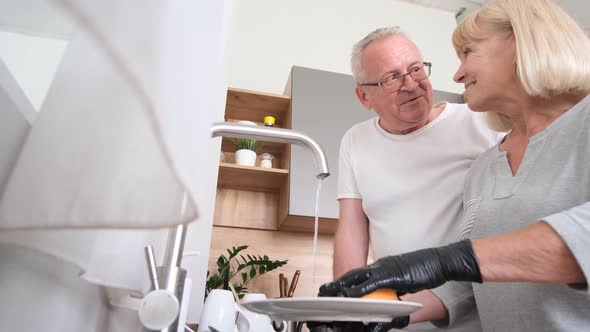 An Elderly Woman is Washing Dishes in the Kitchen and Talking to Her Husband