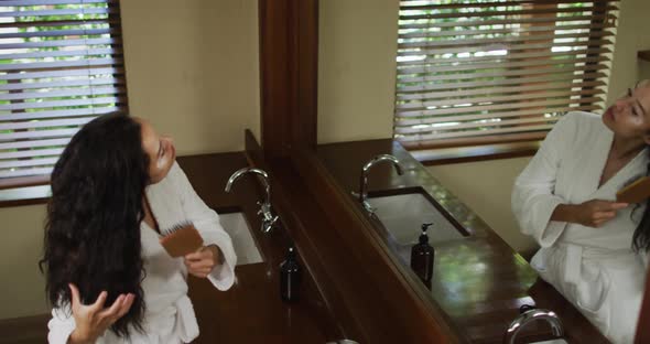 Biracial woman with vitiligo wearing robe looking into mirror and brushing hair in bathroom