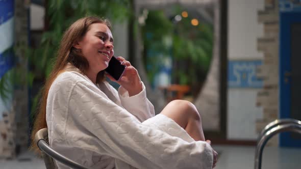 Side View Joyful Millennial Female Tourist Talking on Phone Laughing Sitting at Pool in Hotel