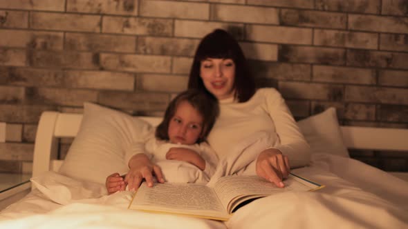 Caucasian Mother Lying in Bed Under the Blanket with Her Daughter Reading Wonderful Fairytales