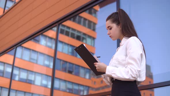 Business Girl with Dark Hair is Making Notes in Working Documents Standing Near Business Center