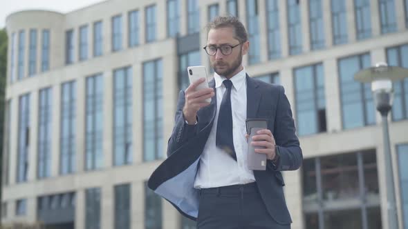Portrait of Concentrated Nerd Businessman Using Smartphone Outdoors in Front of Office Business