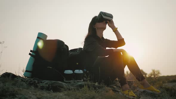 Hipster Young Girl with Backpack on the Top of a Rock Uses Virtual Reality Glasses in the Sunset