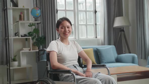 Asian Woman Sitting In A Wheelchair Smiling And Looking At Camera In Living Room