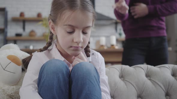 Close-up of Cute Caucasian Brunette Girl with Braided Pigtails Sitting on Sofa and Hugging Knees