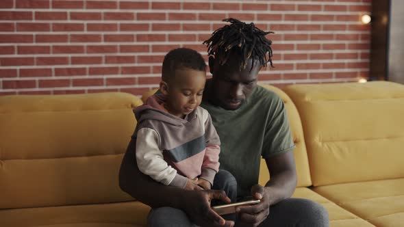 Portrait of African American Father Sitting with Son on Sofa Using Smartphone