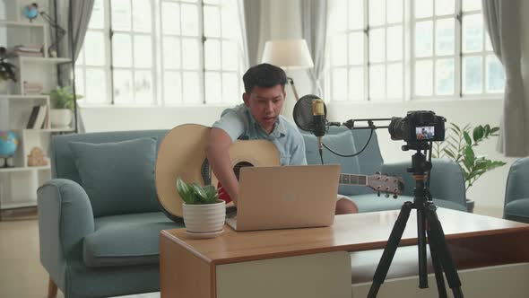 Asian Boy Vlogger With Guitar Looking Comment On Computer Laptop. The Boy Is Streaming