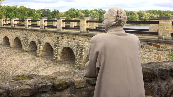 An Elderly Woman Leans Against a Stone Barrier on a Bridge and Looks Around