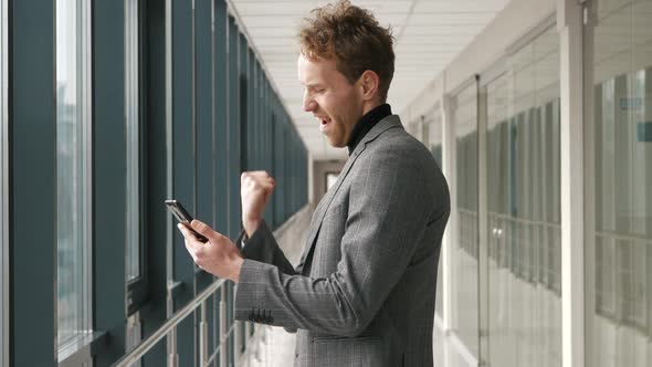 Handsome happy businessman receiving sms message reading good news. Happy male user looking at cellp