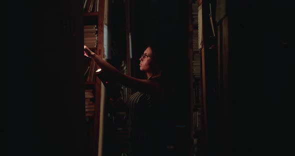 Girl in Glasses with a Lantern Looks for a Book on Library Shelves in Darkness