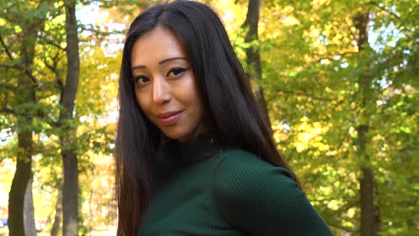 A Young Asian Woman Sits on A Bench in A Park and Smiles at The Camera