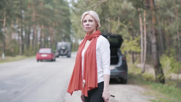 Side View of Adult Blond Woman Standing Next To Broken Car with Wrench and Waiting for Tow Truck