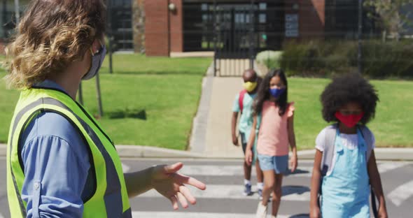 Female woman wearing hi vis vest helping group of kids wearing face masks to cross the road