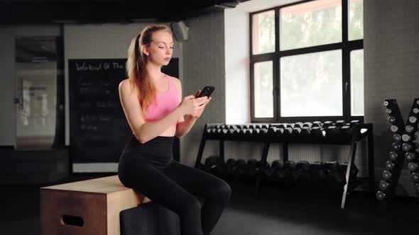 Athlete girl is resting after exercise with the phone while sitting in the gym. Woman texting