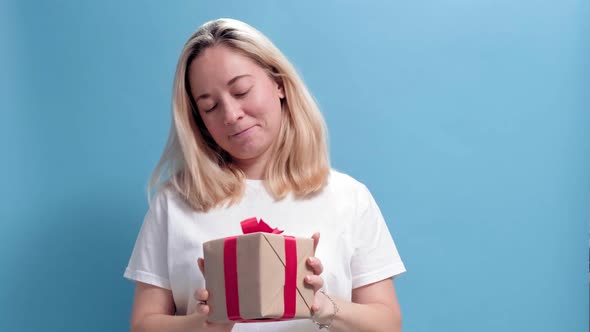 Young Smiling Woman Holding Gift Box and Gives It By Hands to Camera on Blue