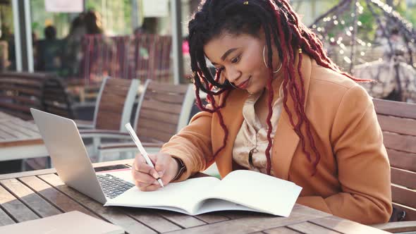 Young African American Woman Sitting in Outddor Cafe with Laptop and Making Notes in Notepad