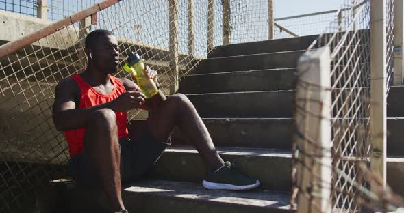 Tired african american man sitting, drinking from water bottle, taking break in exercise outdoors