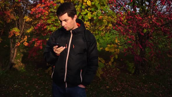 Young Man in Black Jacket in the Autumn Park Using His Smartphone