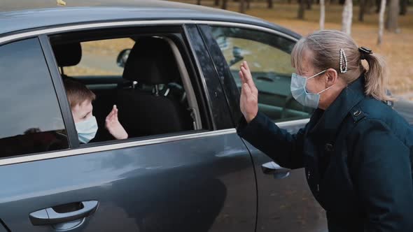 Grandson Boy in a Medical Mask Waving To His Grandmother Through the Car Window. 