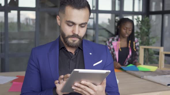 Caucasian Man Looking Pictures on Tablet in the Foreground While African American Female Designer