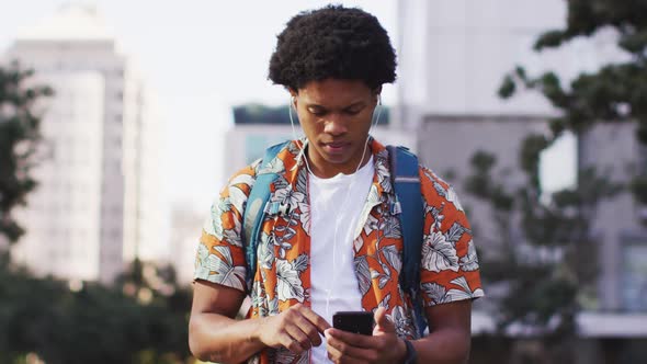 Happy african american man in city, wearing earphones, using smartphone and laughing in the street