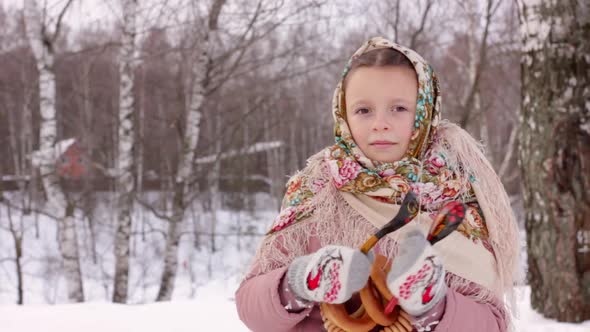 Cute girl in a traditional Russian headscarf and mittens playing on spoons on winter forest backgrou
