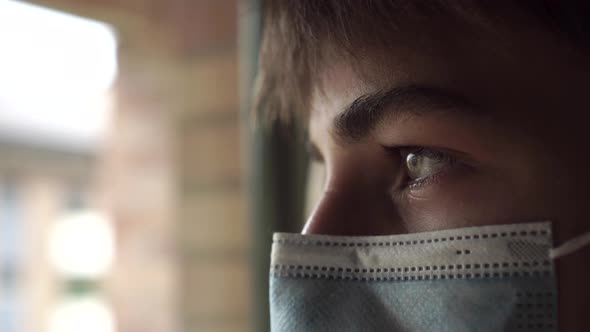 Young man is sad in a medical mask behind a closed window.