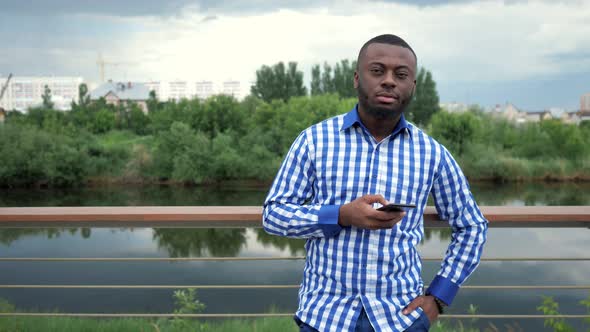 Black Man Browses Phone Looks at Camera and Smiles Stands on Waterfront in Park