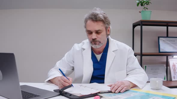 Doctor Sits at the Desk and Talks to Camera Wearing Medical Gown