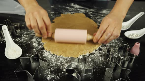 Hands are rolling raw dough with rolling pin on black wood table in modern kitchen