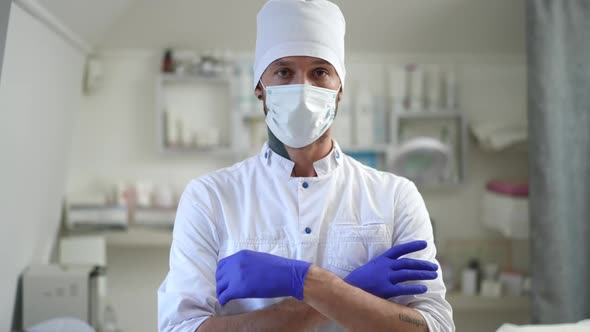 Medium Shot Portrait of Confident Serious Cosmetologist in Covid Face Mask Crossing Hands Looking at