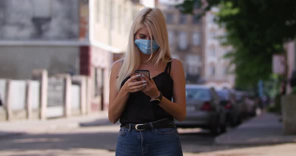 Young Blonde Woman in Protective Medical Mask Walks Down To the Street Uses Phone Texts Scrolls