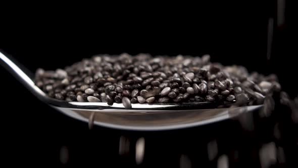 Chia seeds fall into a full metal spoon close up on a black background