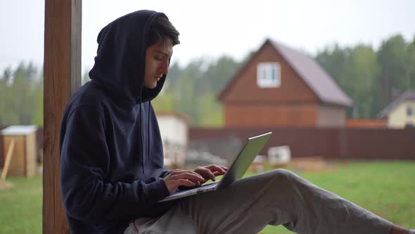 A Young Man is Typing on a Laptop While Sitting on a Terrace Against the Backdrop of a Picturesque