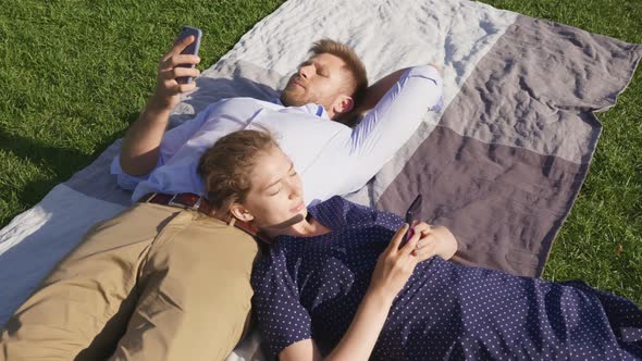 Top View of Young Couple Lying on Blanket in Park Using Cellphone