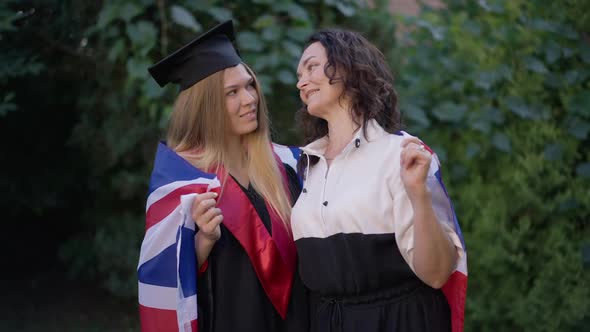 Happy Mature Mother and Young Daughter Wrapped in British Flag Posing Outdoors on Graduation Day