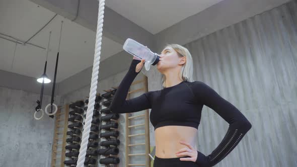 A Young Athletic Blonde Woman Drinks Water from a Bottle After An Exercise Dressed in a Black Top