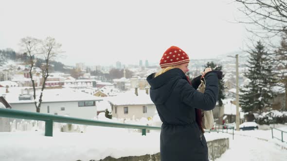 Blonde woman wearing red bonnet and blue coat smiling while taking pictures of snowy Sarajevo