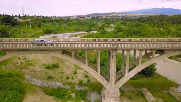 Blue car crosses river on old abandoned bridge against backdrop of mountains