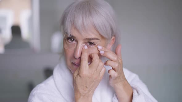 Front View Face of Mature Caucasian Woman Applying Eye Pads Looking at Camera Standing in Bathroom