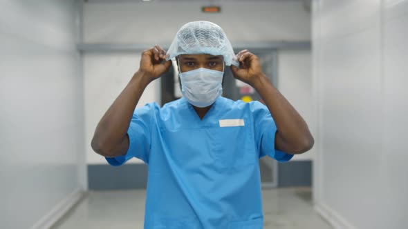 Young Male Afro Doctor Wearing Scrubs and Cap Walking Along Hospital Corridor