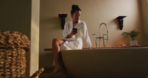 Biracial woman sitting in robe on bathtube, preparing bath, using smartphone